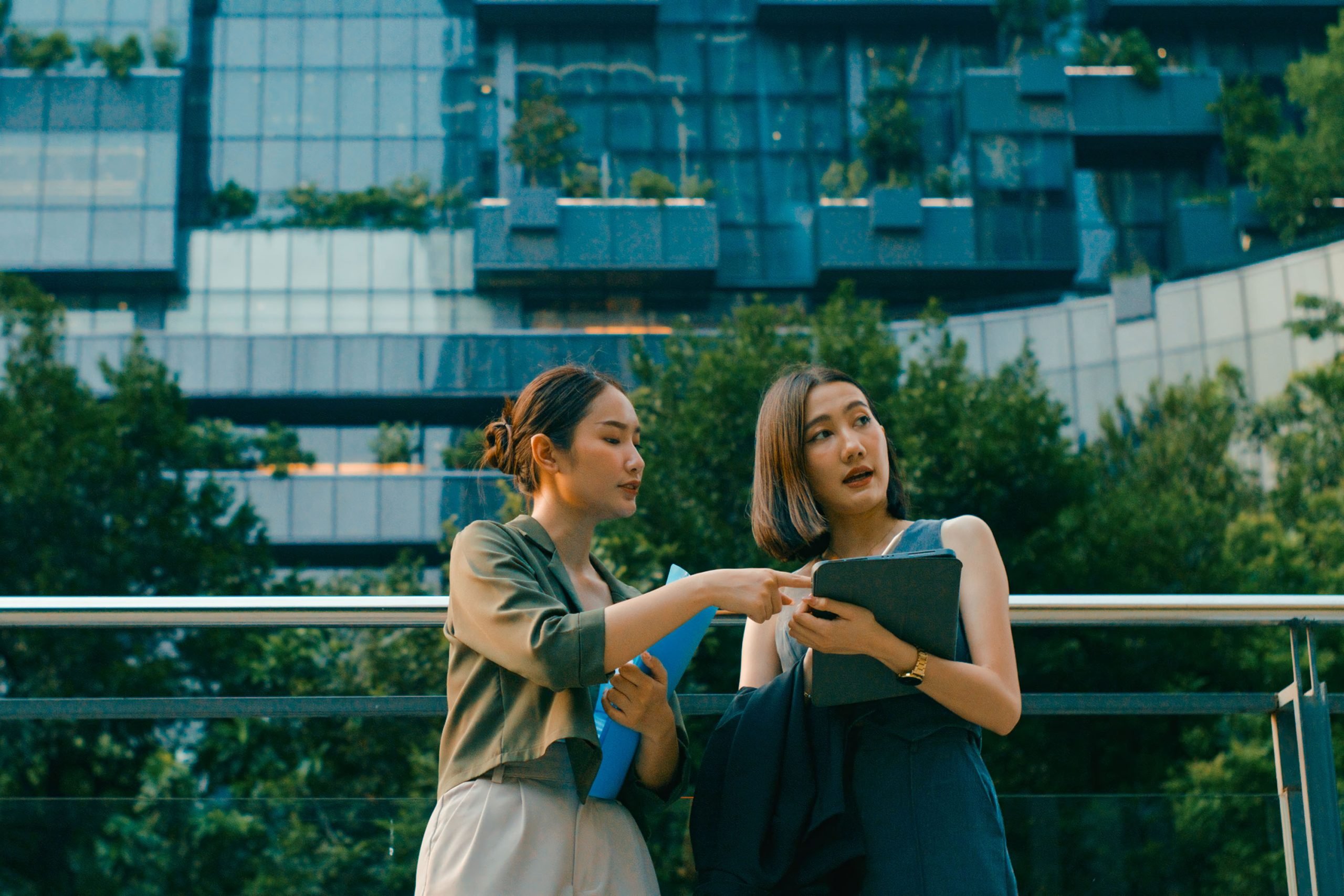 Two young Asian businesswomen meeting business strategy discussion talk outside modern office building. Sustainable business outdoor.